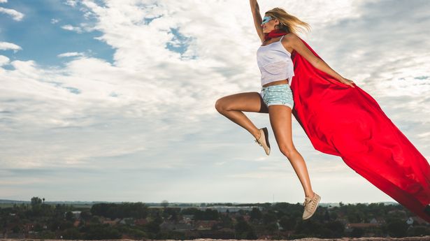 Blonde woman in red dress and red mantle jumping outdoor as a superhero against blue sky